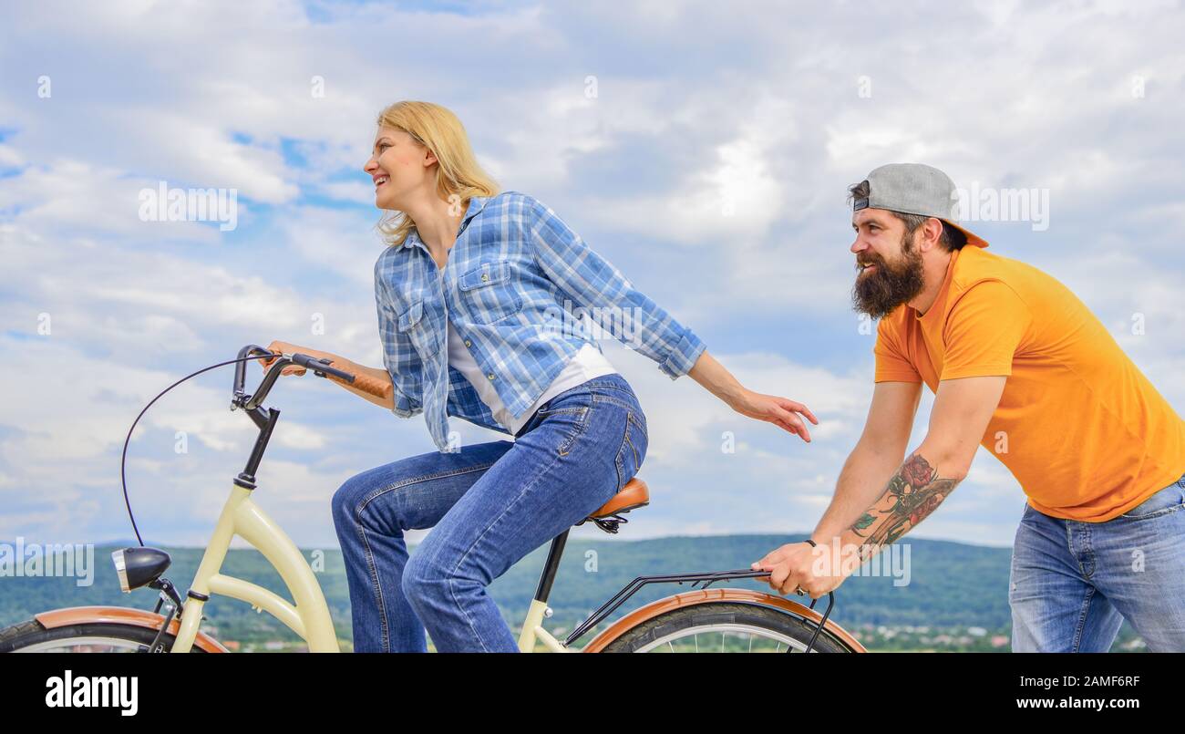 Vélo fille tandis que l'homme la soutenir. Aide à croire en vous-même. Impulsion L'impression de commencer à bouger. Des promenades en vélo femme fond de ciel. Pousser et la promotion. Impulsion à se déplacer. Homme pousse fille faire du vélo. Banque D'Images