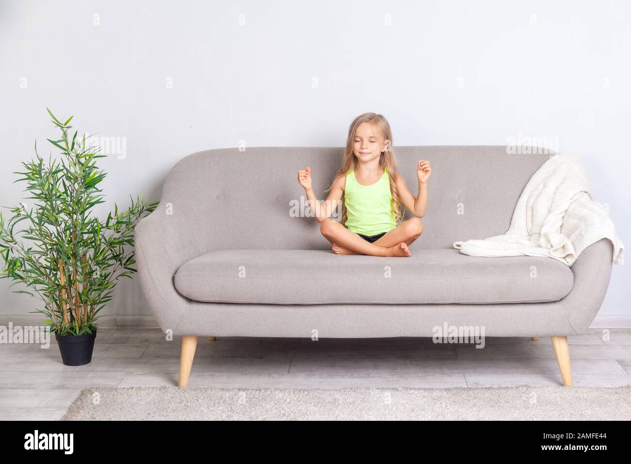 Jolie petite fille calme méditant dans lotus pose avec des yeux fermés assis sur le canapé à la maison, enfant faisant le geste de mudra, paisible heureux expression visage, h Banque D'Images