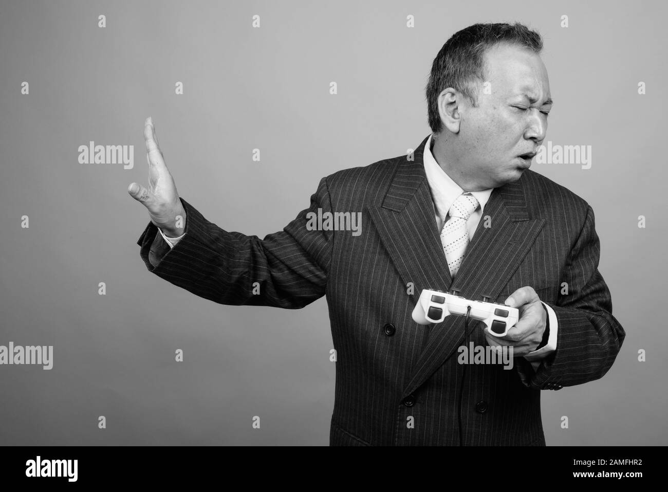 Studio tourné d'un homme d'affaires asiatique mûr jouant à des jeux sur fond gris en noir et blanc Banque D'Images Studio tourné d'un homme d'affaires asiatique mûr jouant à des jeux sur fond gris en noir et blanc Banque D'Images