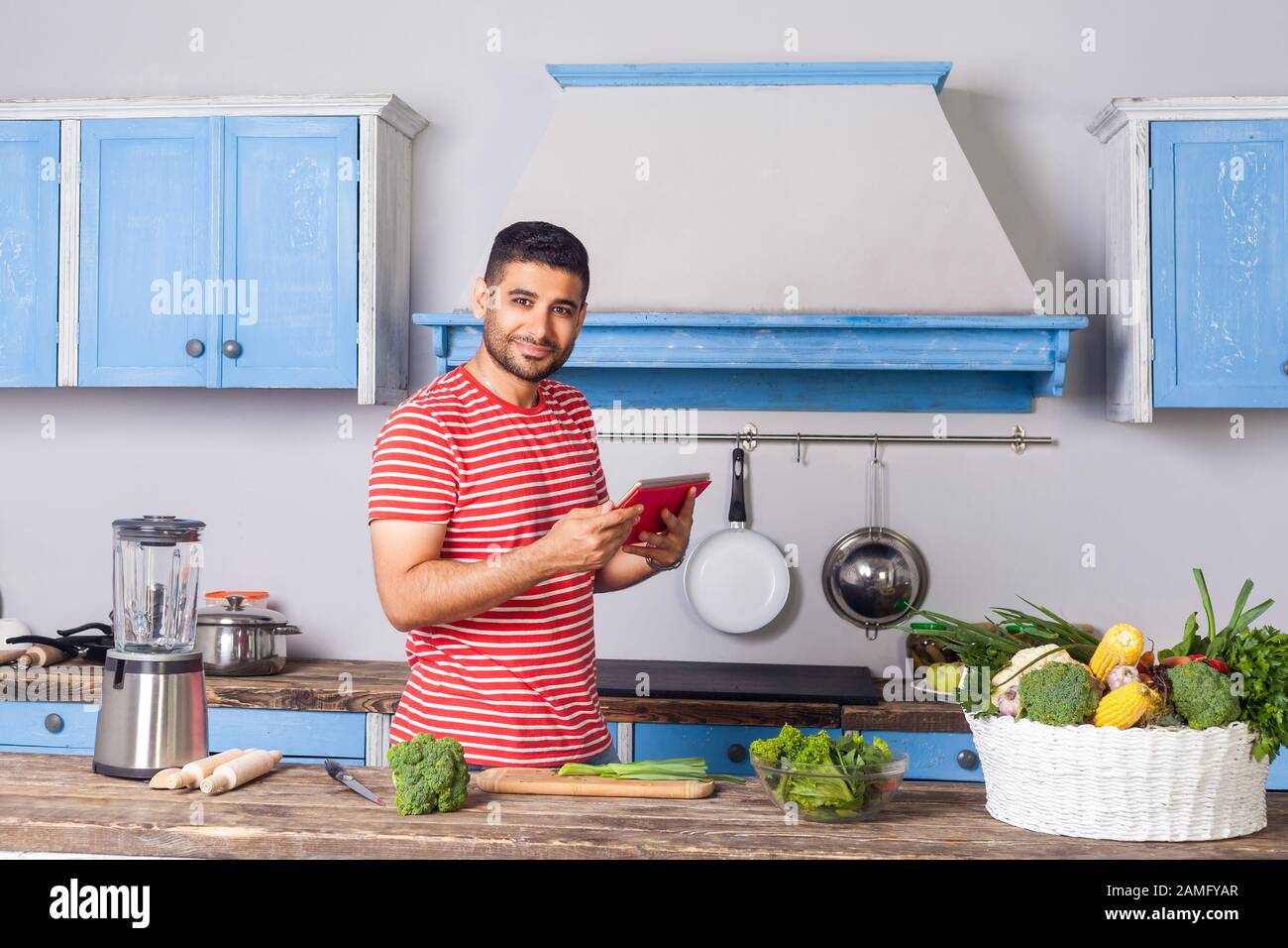 Homme positif debout dans la cuisine moderne tenant tablette pc et regardant l'appareil photo avec le sourire, la recherche de la recette de nourriture sur Internet, la lecture des calories, baske Banque D'Images