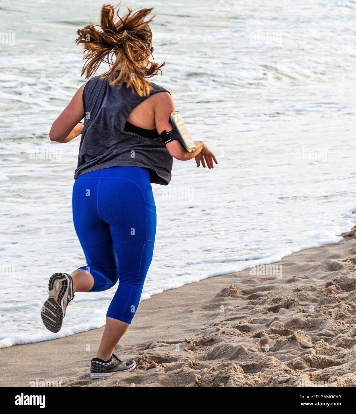 Vue arrière d'un serveur en spandex bleu qui tourne à côté de l'eau sur le sable avec un téléphone portable attaché à son bras pour écouter de la musique. Banque D'Images
