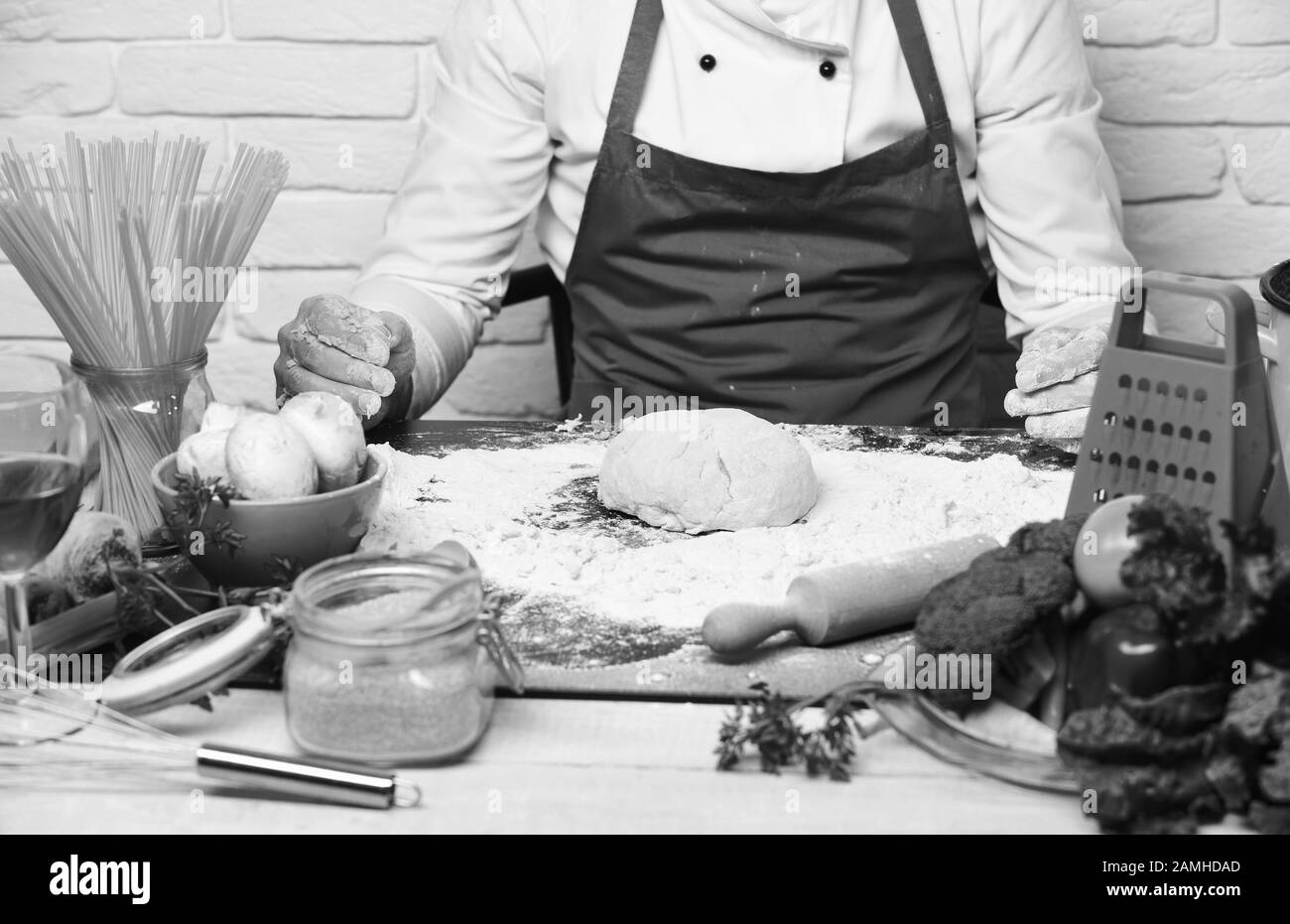 Concept du processus de cuisson. Les mains des hommes travaillent avec de la pâte à pétrir sur fond blanc de mur de brique, gros plan. Faites cuire en Bourgogne en uniforme assis à la table avec des légumes et des ustensiles de cuisine. Le chef fait de la pâte. Banque D'Images