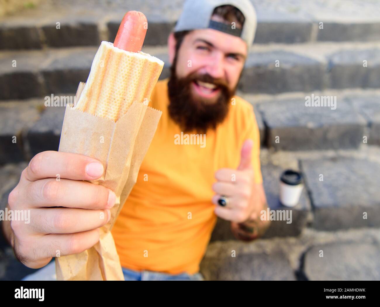 Homme barbu profiter de la nourriture de rue escalier arrière-plan. Hipster mange un chien chaud et boit du café. Concept de l'heure du déjeuner. Hipster appréciez la boisson chaude de votre chien et de votre tasse en papier. Restauration rapide pour déjeuner idée commune. Banque D'Images