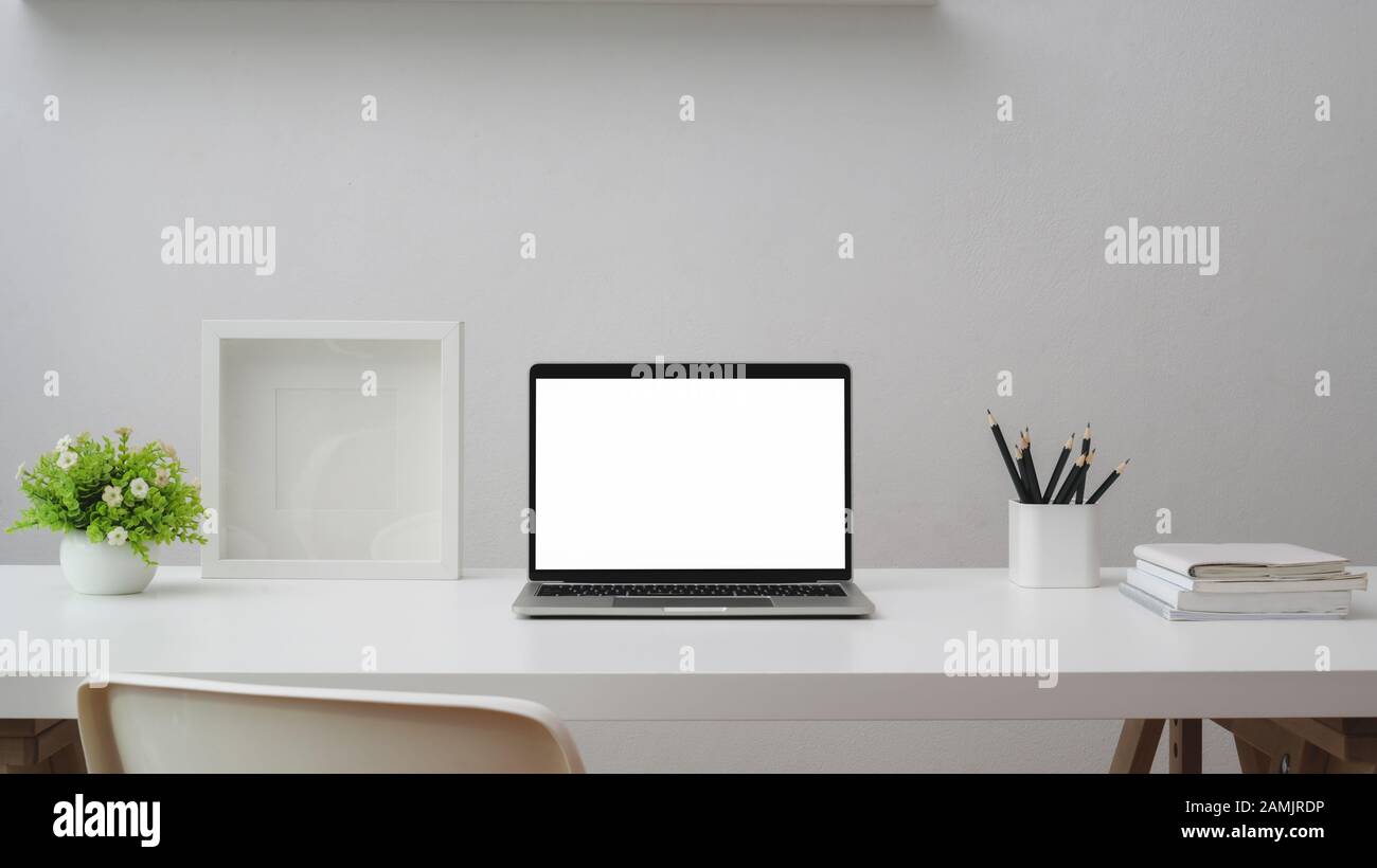 Gros plan sur un espace de travail moderne avec un ordinateur portable à écran vide, un cadre, des articles de papeterie et des pots d'arbres sur une table blanche avec fond de mur blanc Banque D'Images