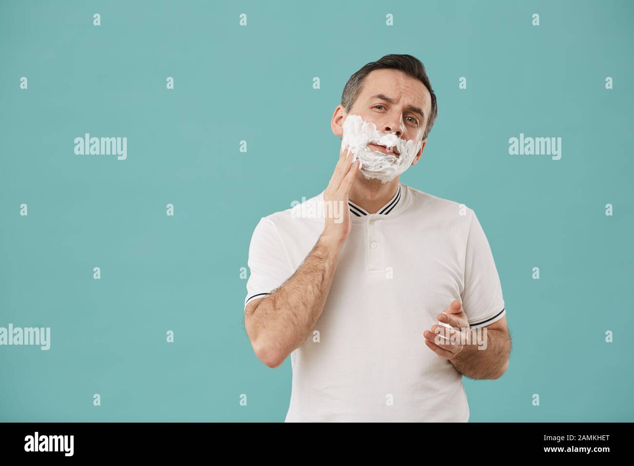 Taille relevée portrait de l'homme moderne d'âge moyen rasage et regardant l'appareil photo tout en appréciant la routine du matin debout contre fond bleu, espace de copie Banque D'Images