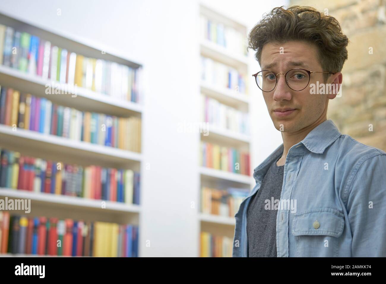 Jeune étudiant avec des boucles et des verres, à la recherche sans heurts et surchargés dans l'appareil photo, dans la bibliothèque, à l'université. Banque D'Images