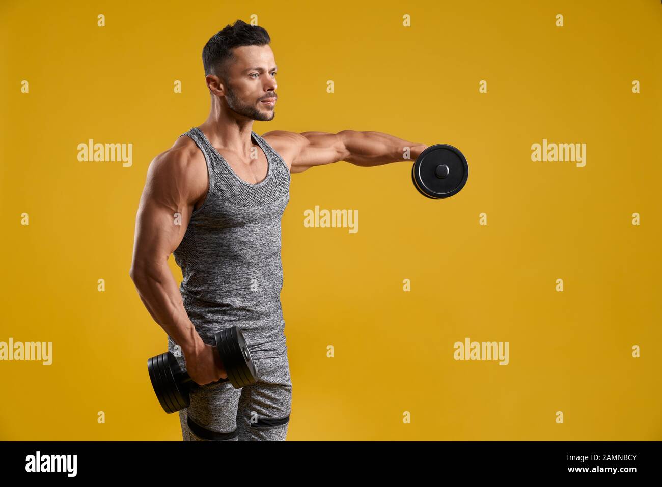 Vue latérale d'un beau fer de pompage bodybuilder. Mec musclé de soulever des poids lourds et d'haltères à côté avec sérieux. Jaune isolé sur fond de studio. Concept d'entraînement intensif. Banque D'Images