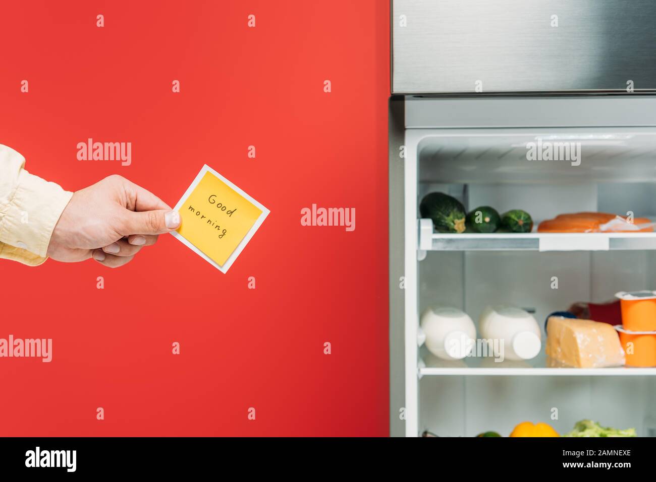vue réduite de l'homme tenant un pense-bête avec un bon lettrage le matin près d'un réfrigérateur ouvert avec des aliments frais sur des étagères isolées en rouge Banque D'Images