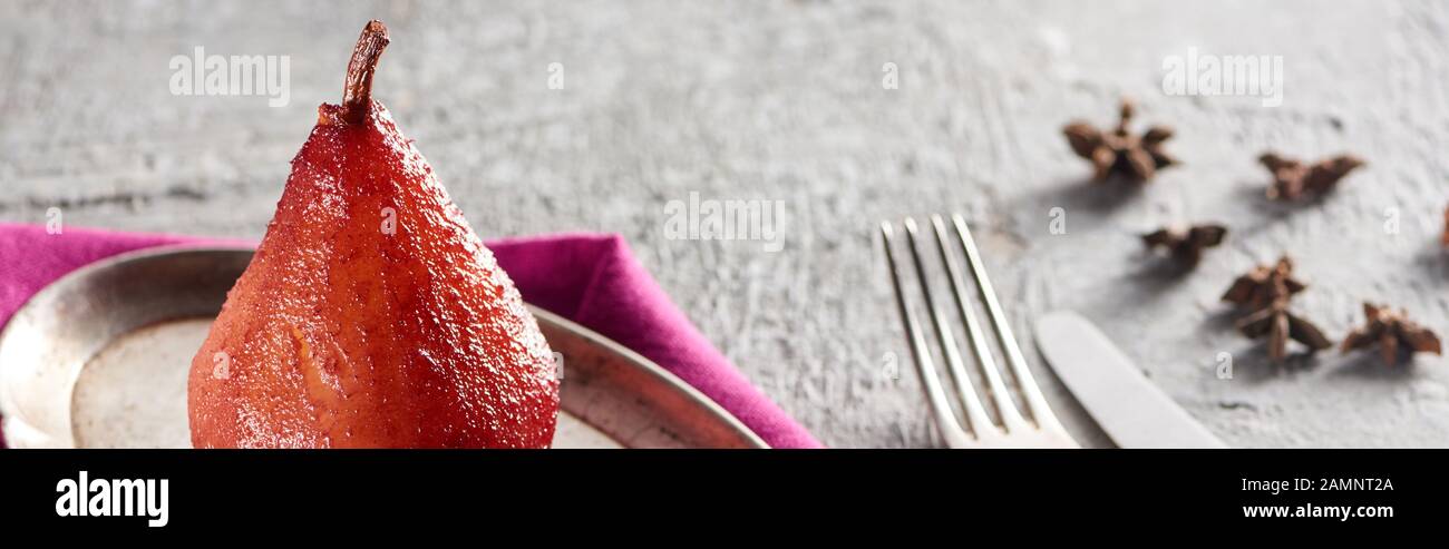 délicieuse poire en vin avec anis sur plaque argentée sur surface en béton gris avec serviette rose, couteau et fourchette, vue panoramique Banque D'Images