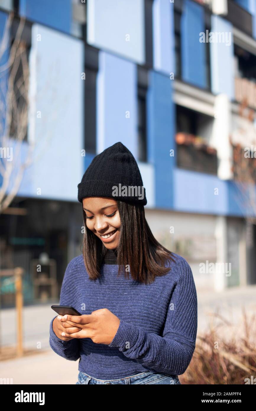 Portrait de la jeune femme africaine américaine portant une casquette en laine debout dans la rue tout en utilisant un téléphone portable Banque D'Images