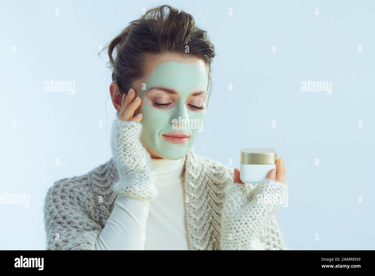 femme élégante de 40 ans, sweat-shirt à col roulé et gilet avec masque facial vert montrant un pot cosmétique sur fond bleu clair d'hiver. Banque D'Images