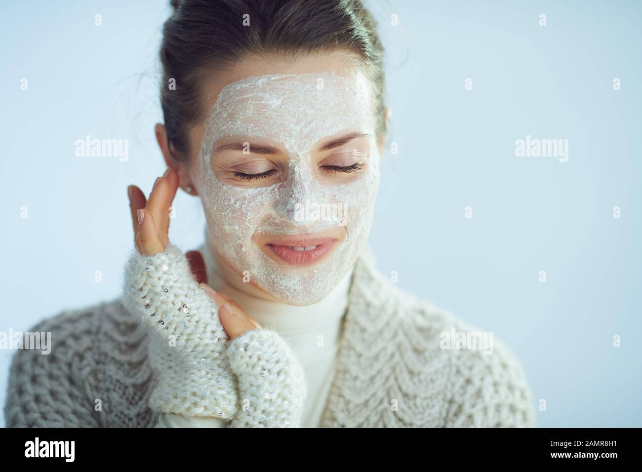 femme moderne et décontractée de 40 ans, sueur à col roulé et gilet avec masque facial blanc dans le cadre des soins de la peau d'hiver isolés sur bleu clair d'hiver b Banque D'Images