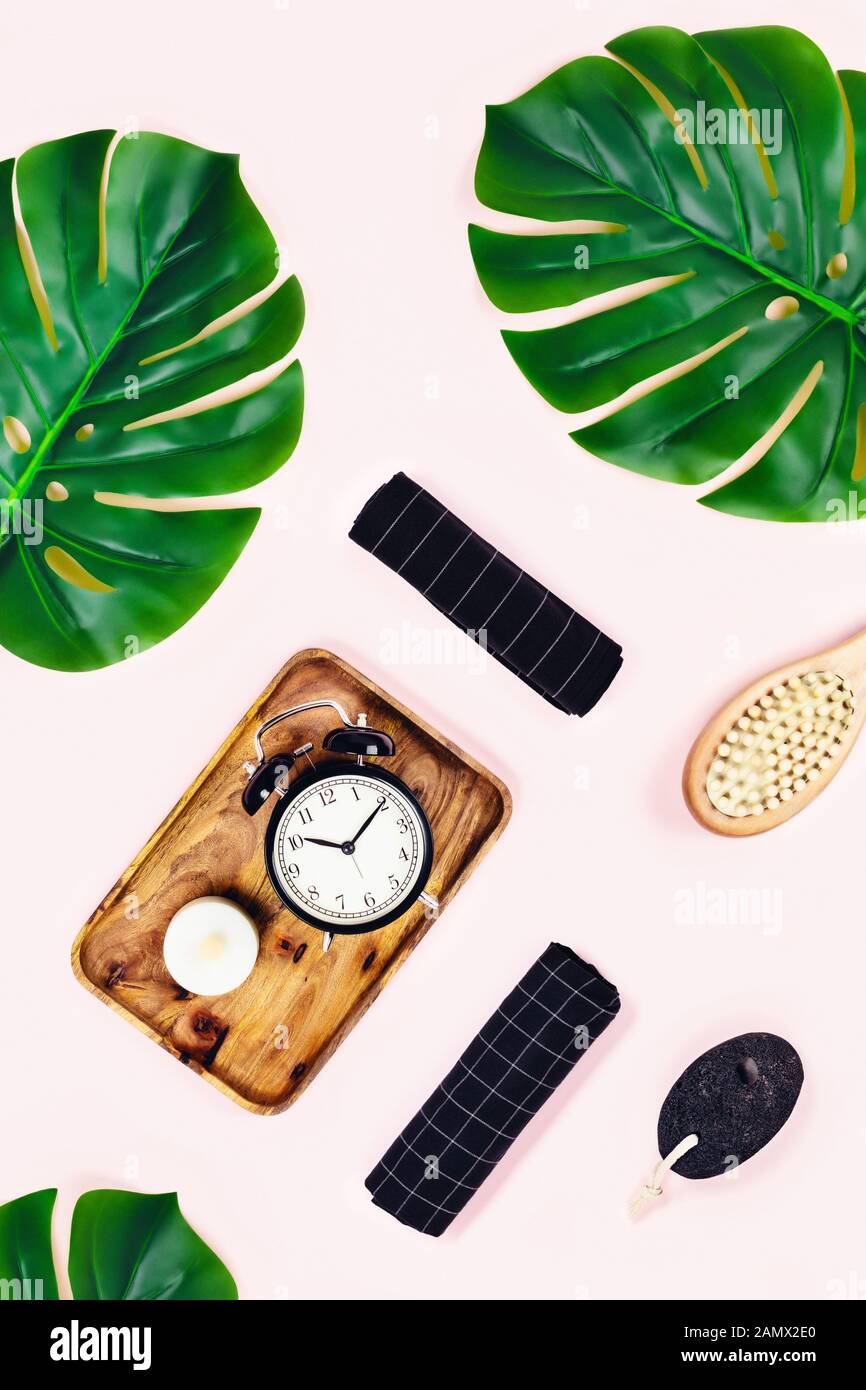 Fond rose avec plateau en bois et réveil noir, pierre ponce entourée de feuilles tropicales. Plat, vue du dessus. Concept de spa. Banque D'Images