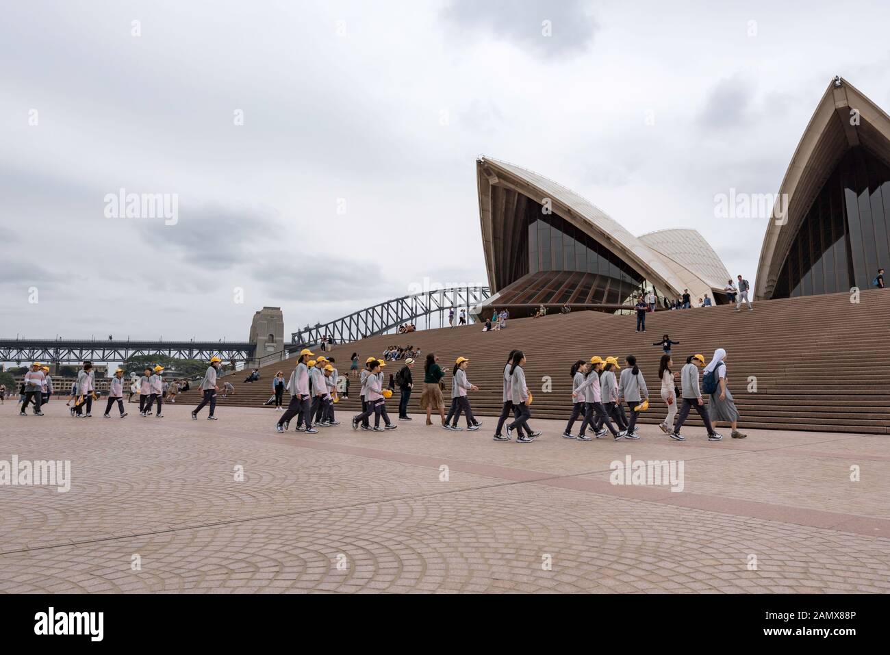 Un nun catholique dirige un groupe de jeunes enfants asiatiques âgés d'une école primaire, portant principalement des casquettes jaunes, vers les marches de l'Opéra de Sydney. Banque D'Images