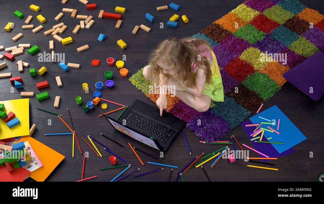 Un enfant d'âge préscolaire blond et corné regarde un dessin animé sur un ordinateur portable, assis au sol Banque D'Images