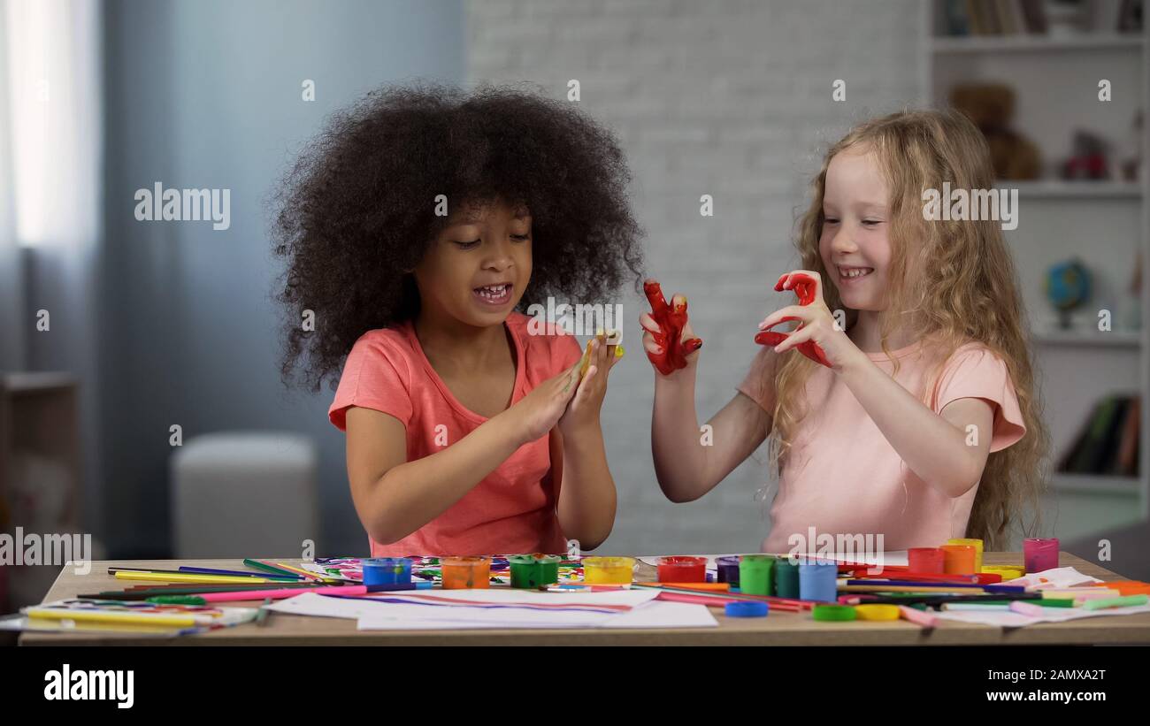 Filles multiethniques au club pour enfants, assis à la table et se moquant des mains avec des peintures Banque D'Images