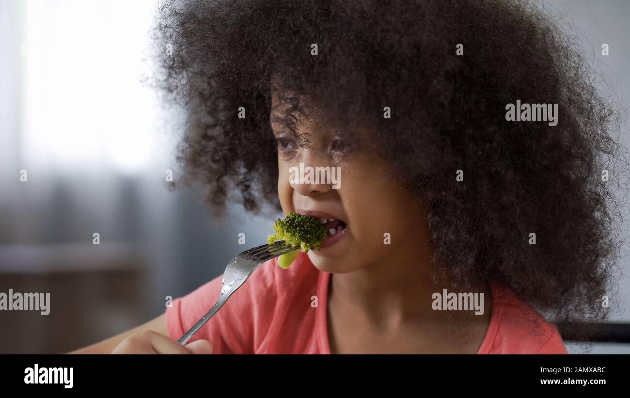 Mignonne jeune fille africaine qui mange du brocoli avec dégoût, nourriture saine, régime Banque D'Images