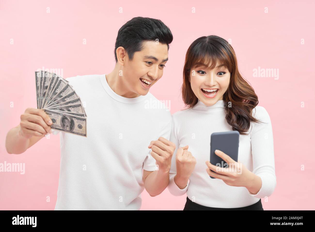 Image d'un jeune couple asiatique gai isolé sur fond de mur rose tenant de l'argent et téléphone mobile. Banque D'Images