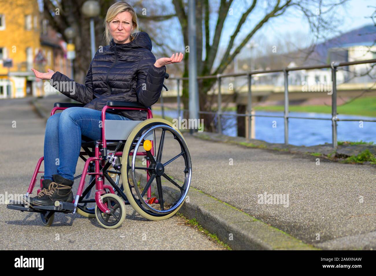 Femme handicapée dans un fauteuil roulant en haussant les épaules avec un sourire ironique qu'elle siège en attente dans une rue urbaine calme le long d'une rivière Banque D'Images