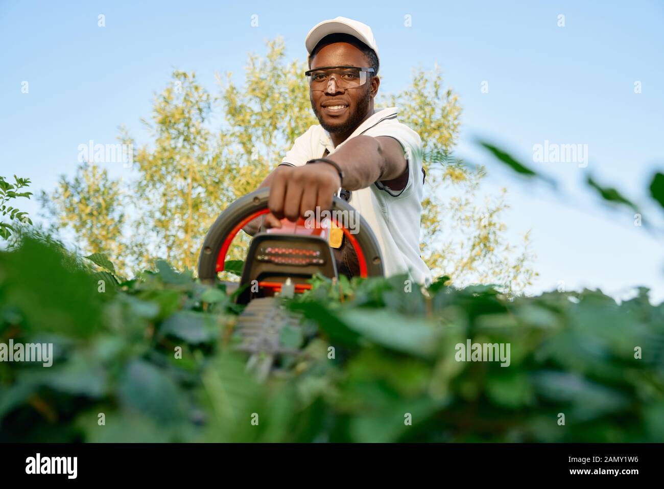Portrait of smiling african man in lunettes de protection et d'élagage hat haie avec tondeuse à main. Homme jardinier dans vêtements de travail réduisant les buissons pendant les beaux jours. Banque D'Images