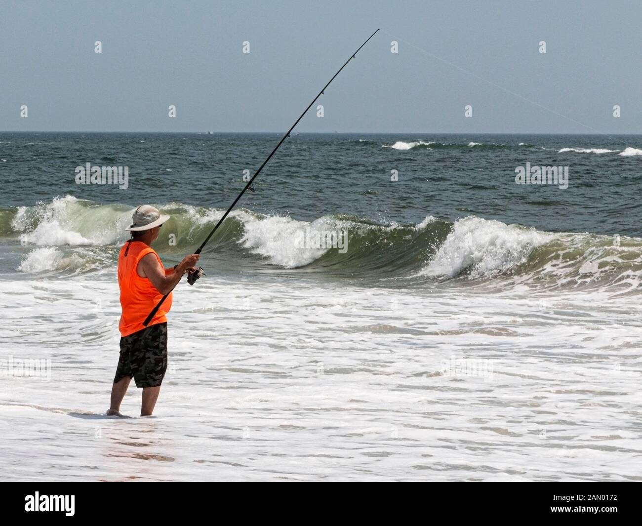 Un homme dans une chemise orange est la pêche de surf coulés dans l'océan avec de petites vagues se brisant devant lui. Banque D'Images
