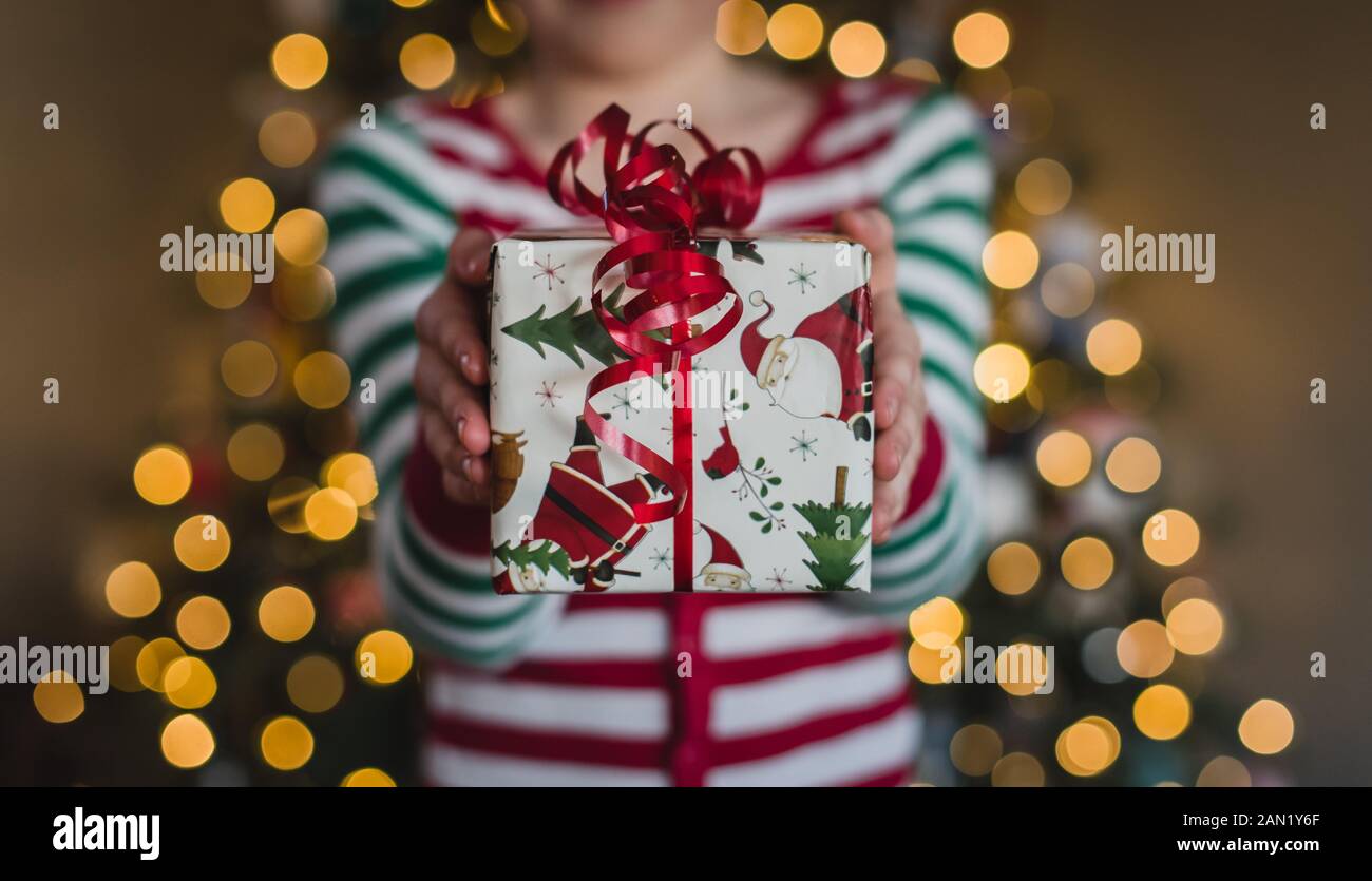 Enfant en pyjama tenant un cadeau de Noël devant les lumières de l'arbre. Banque D'Images