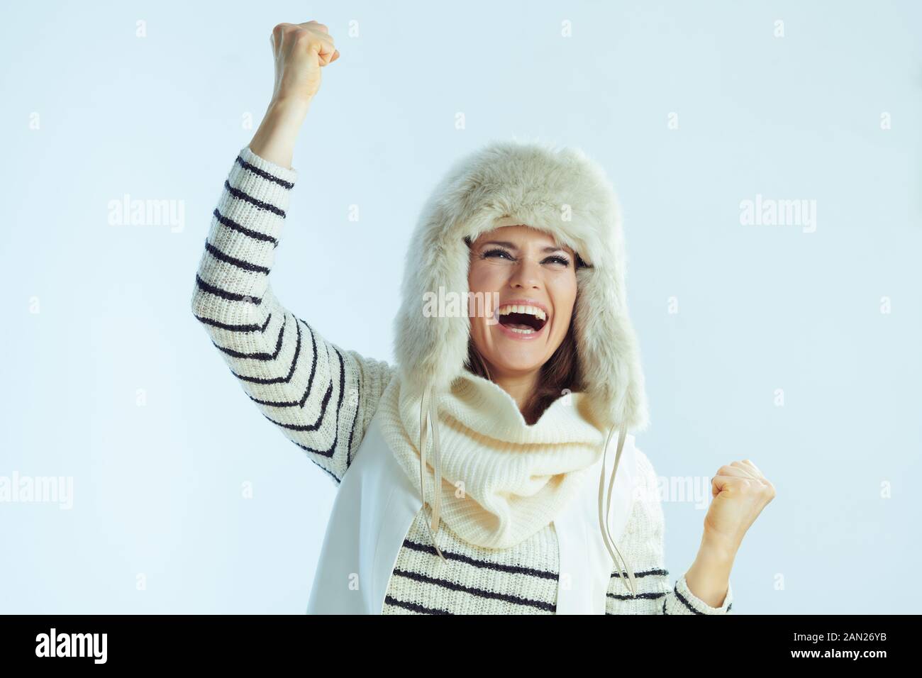 femme souriante et élégante de 40 ans, sweat à rayures blanches, écharpe et rabats pour les oreilles chapeau se réjouissant sur fond bleu clair d'hiver. Banque D'Images