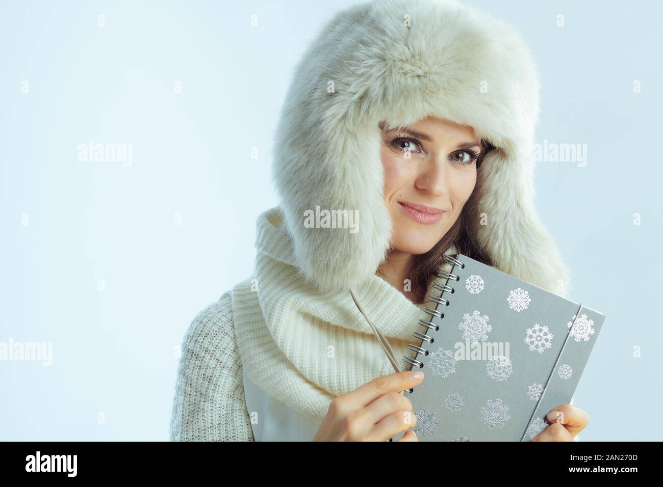 Portrait d'une femme moderne souriante de 40 ans en sweat-shirt blanc à rayures, écharpe et rabats pour les oreilles chapeau tenant le carnet contre fond bleu clair d'hiver. Banque D'Images