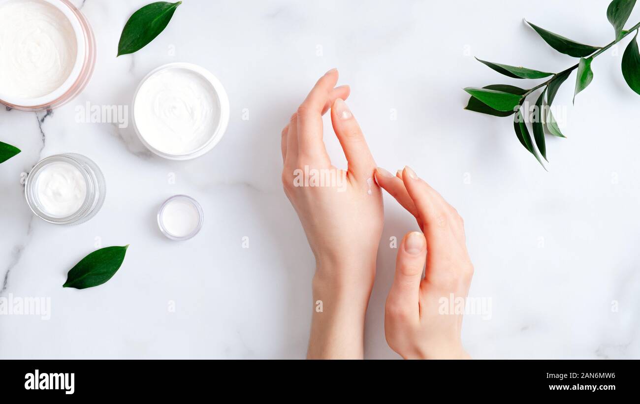 Crème cosmétique sur les mains, pots avec du lait et crème swirl vert feuilles sur la table de marbre blanc. Mise à plat, vue du dessus. L'application femme moisturi organique Banque D'Images