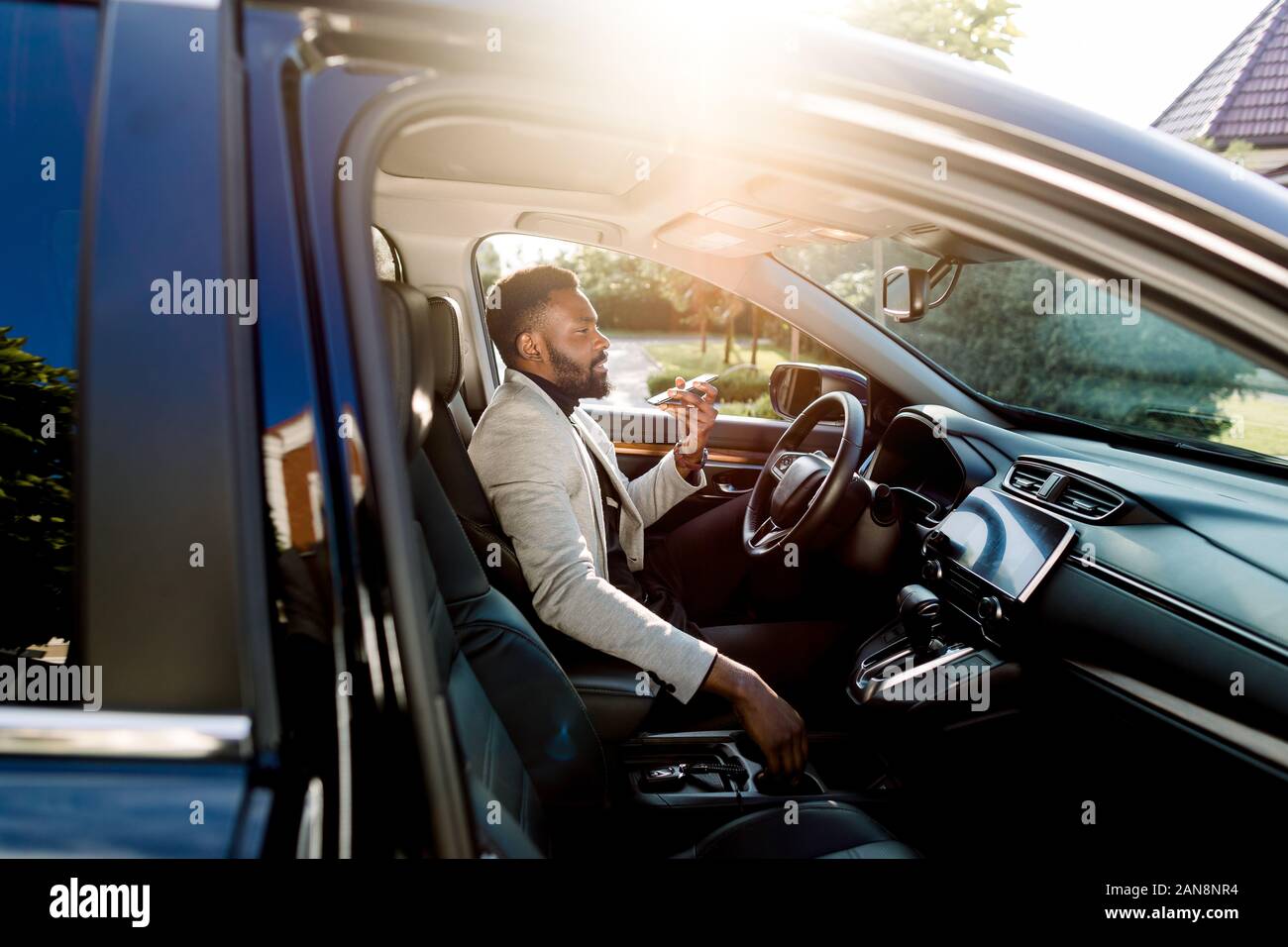 Young handsome African businessman using mobile phone, parlant sur le haut-parleur, tout en étant assis à l'intérieur d'une voiture de luxe Banque D'Images
