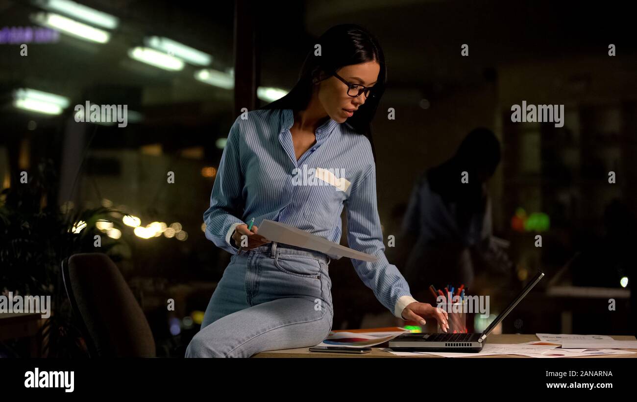 Jeune employé de compagnie avec des papiers la main à l'aide d'un ordinateur portable, travaillant de nuit in office Banque D'Images