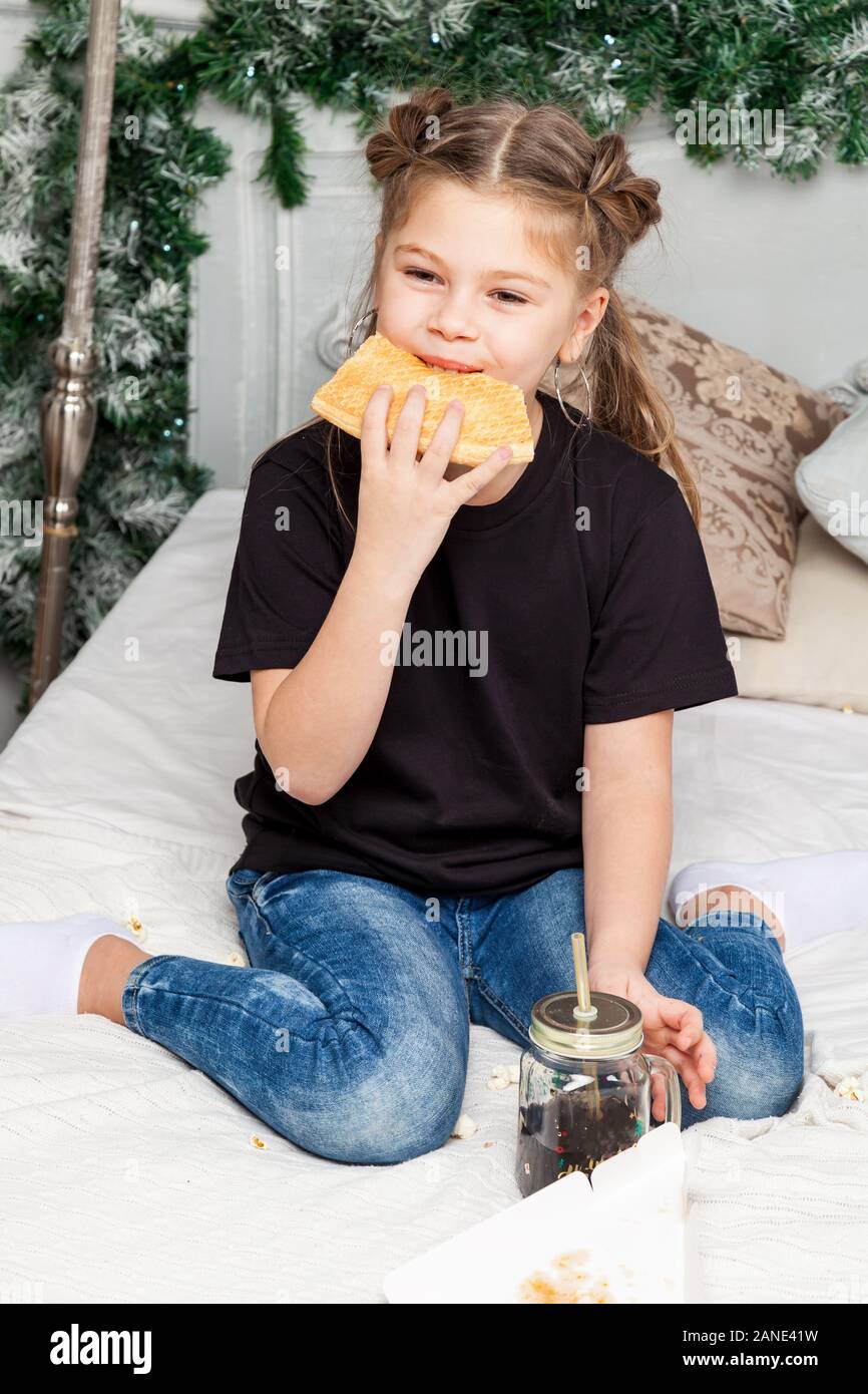 Peu cute belle fille est assise en tailleur sur un lit dans un jean bleu et une chemise noire de manger des pizzas et boire dans le contexte d'une chambre à la N Banque D'Images