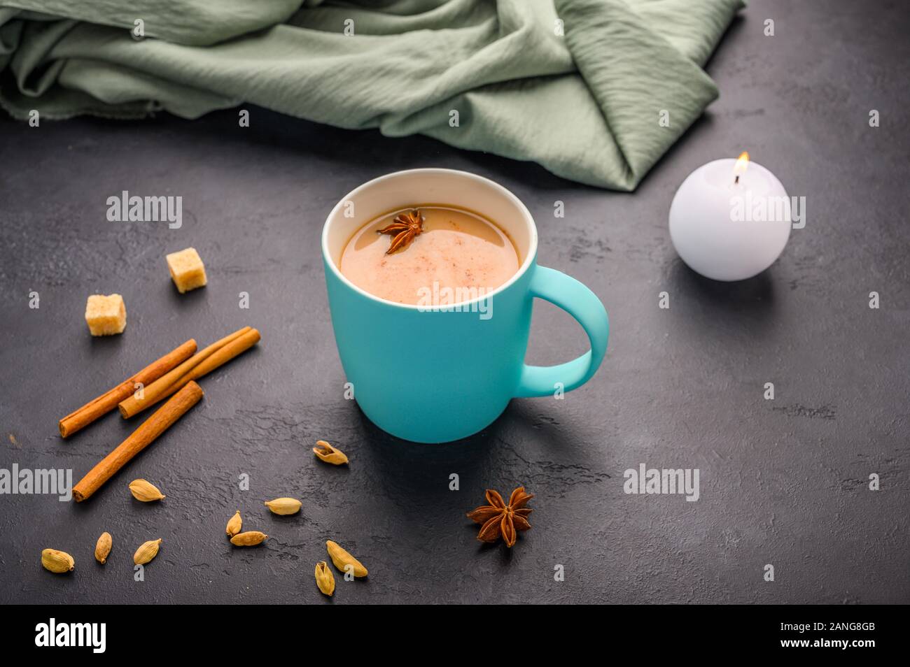 Thé traditionnel indien masala chai dans une tasse bleue. Bougie allumée, la cannelle, l'anis, la cardamome, le sucre à côté d'un fond texturé noir Banque D'Images