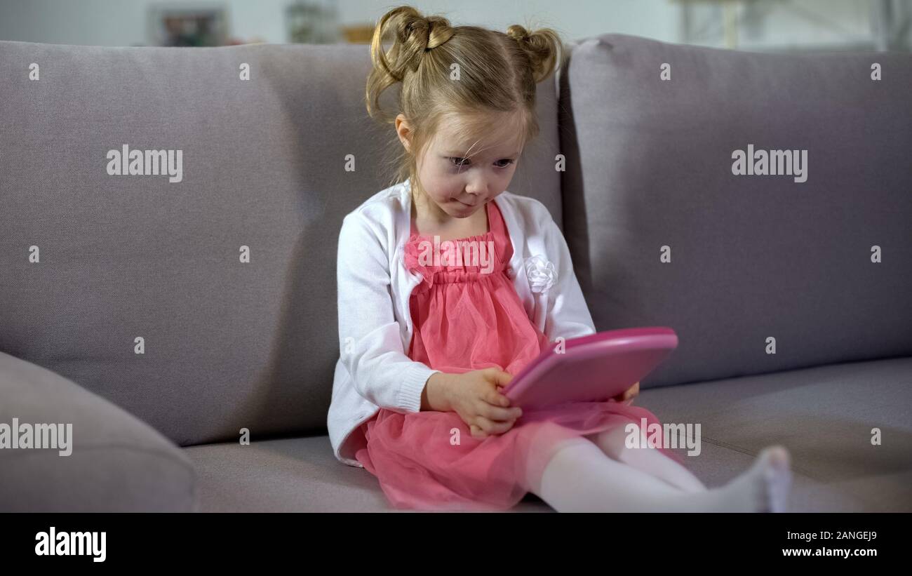 Funny little girl sitting on sofa et jouant sur des jouets éducatifs rose tablet app Banque D'Images