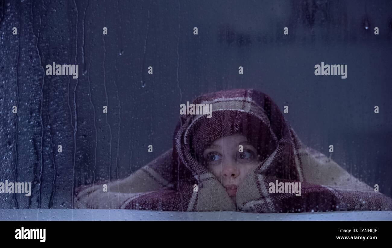 Fille effrayée couvert de couverture sur sill, regardant par la fenêtre la pluie, peurs Banque D'Images