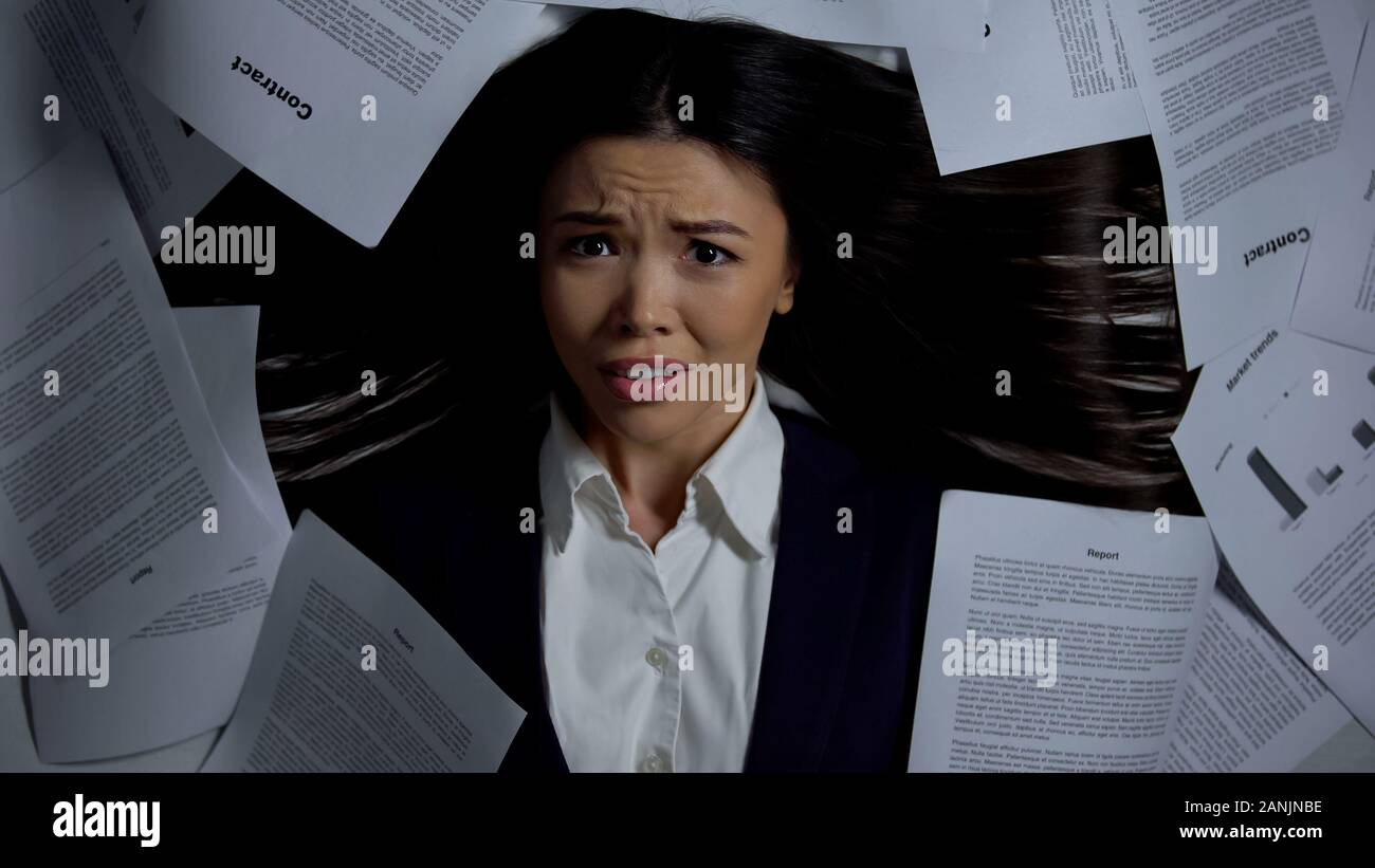 Woman looking at camera, avec horreur, se prélassant parmi les tas de papiers, travail stressant Banque D'Images