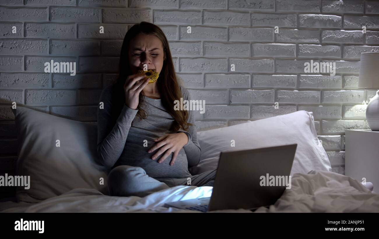 Jeune femme enceinte de pleurer en face de l'ordinateur portable, manger donut de surmonter le stress Banque D'Images