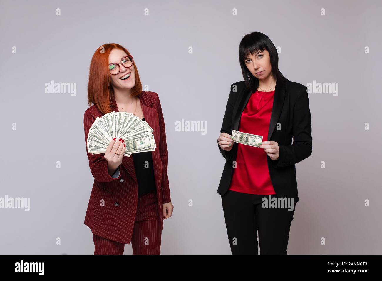 Stock photo portrait of two businesswomen avec de l'argent. Femme aux cheveux rouges en costume rouge tenant une grosse somme de dollars. Femme aux cheveux noirs en costume noir Banque D'Images