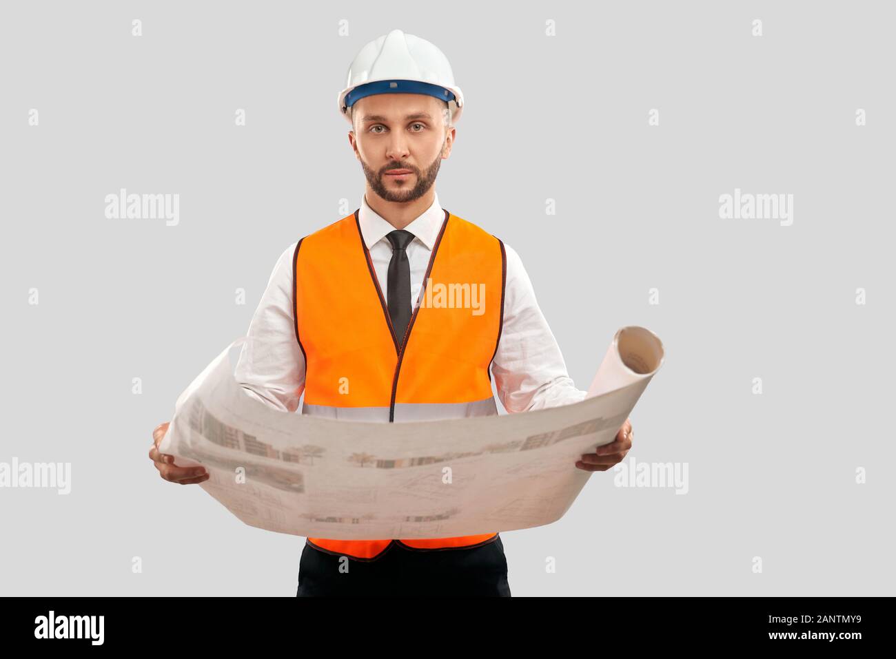 Portrait du jeune architecte dans un gilet réfléchissant orange et un casque tenant une grande carte. Plan de construction en mariage avec un ingénieur, en regardant la caméra, isolé sur gris. Concept d'architecture, de construction. Banque D'Images
