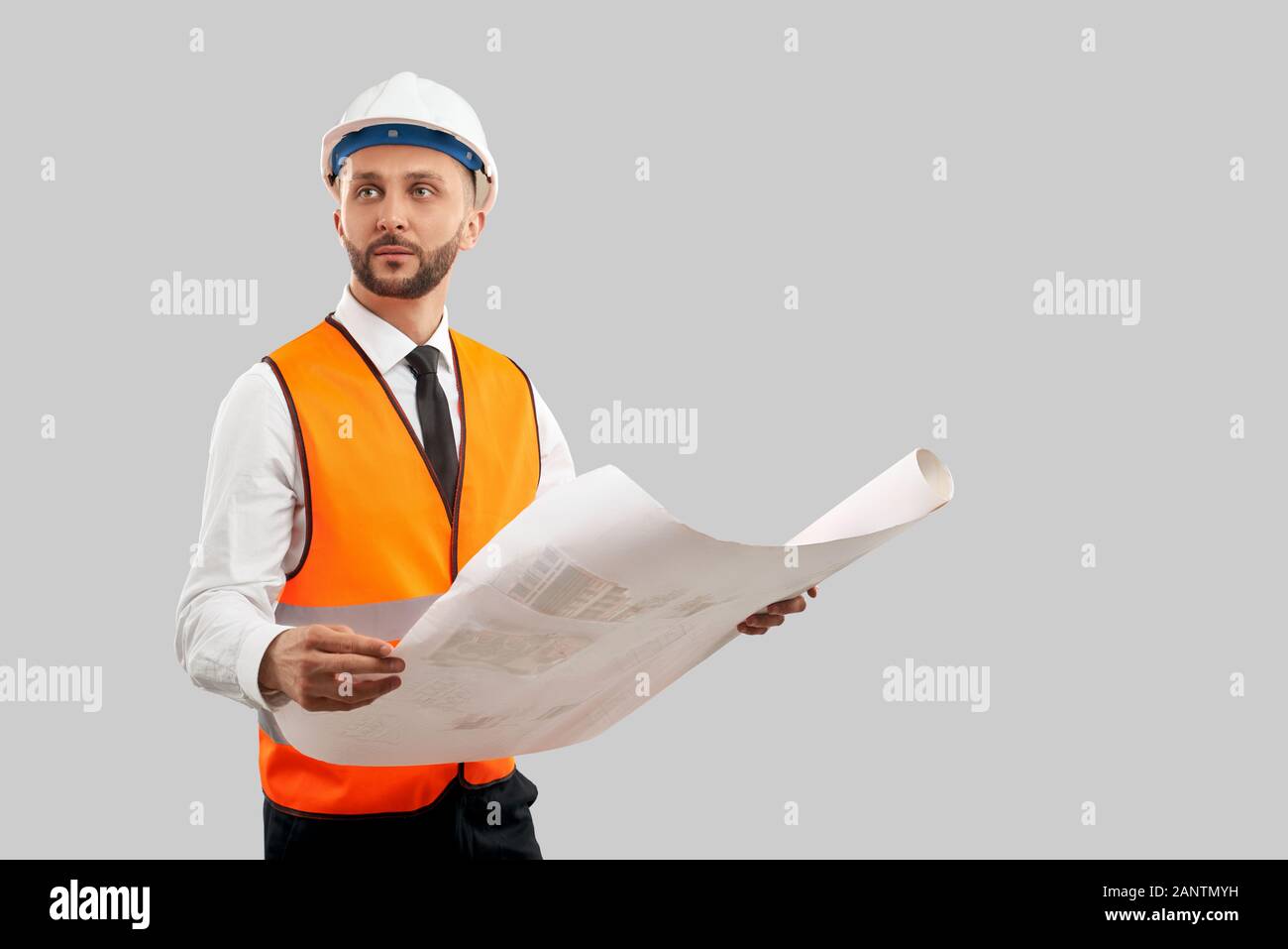 Vue avant du jeune architecte barbu avec un gilet réfléchissant orange et un casque tenant une grande carte. Plan de construction de l'exploitation de carrières, recherche, isolé sur gris. Concept d'architecture, de construction. Banque D'Images