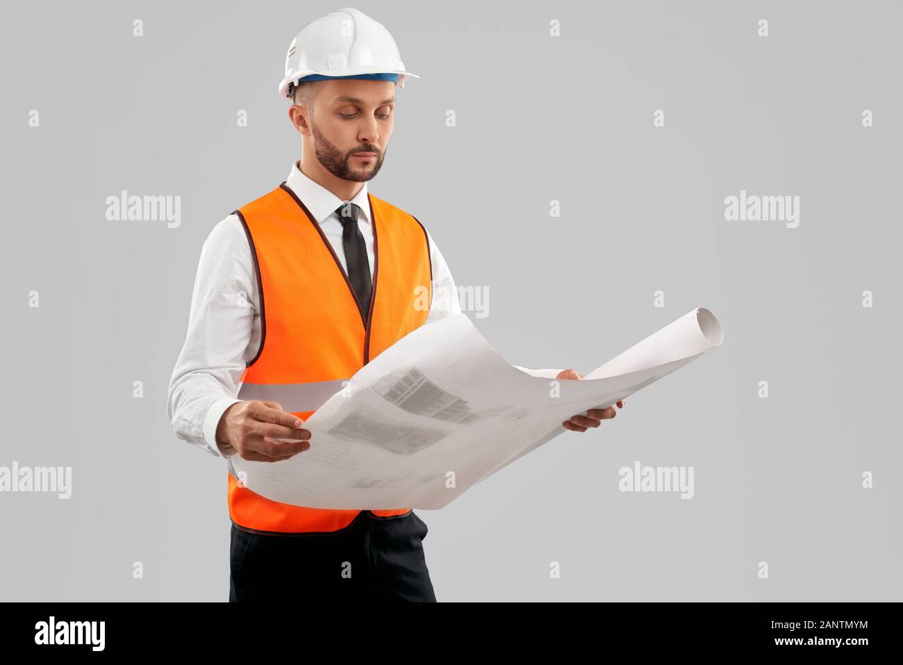 Vue avant du jeune architecte barbu avec un gilet réfléchissant orange et un casque tenant une grande carte. Ingénieur analysant le plan de construction, isolé sur gris. Concept d'architecture, de construction. Banque D'Images
