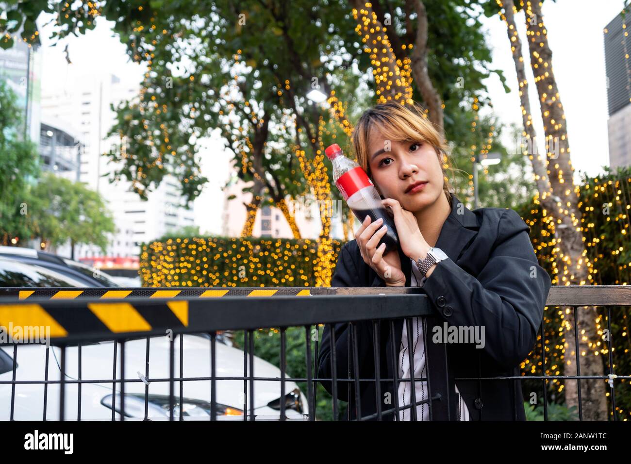 La moitié des cheveux blonds attachés femme de noir se tenant à la barrière noire et se pelotonnant jusqu'à bouteille de boisson dans le stationnement avec décoration lumière Banque D'Images