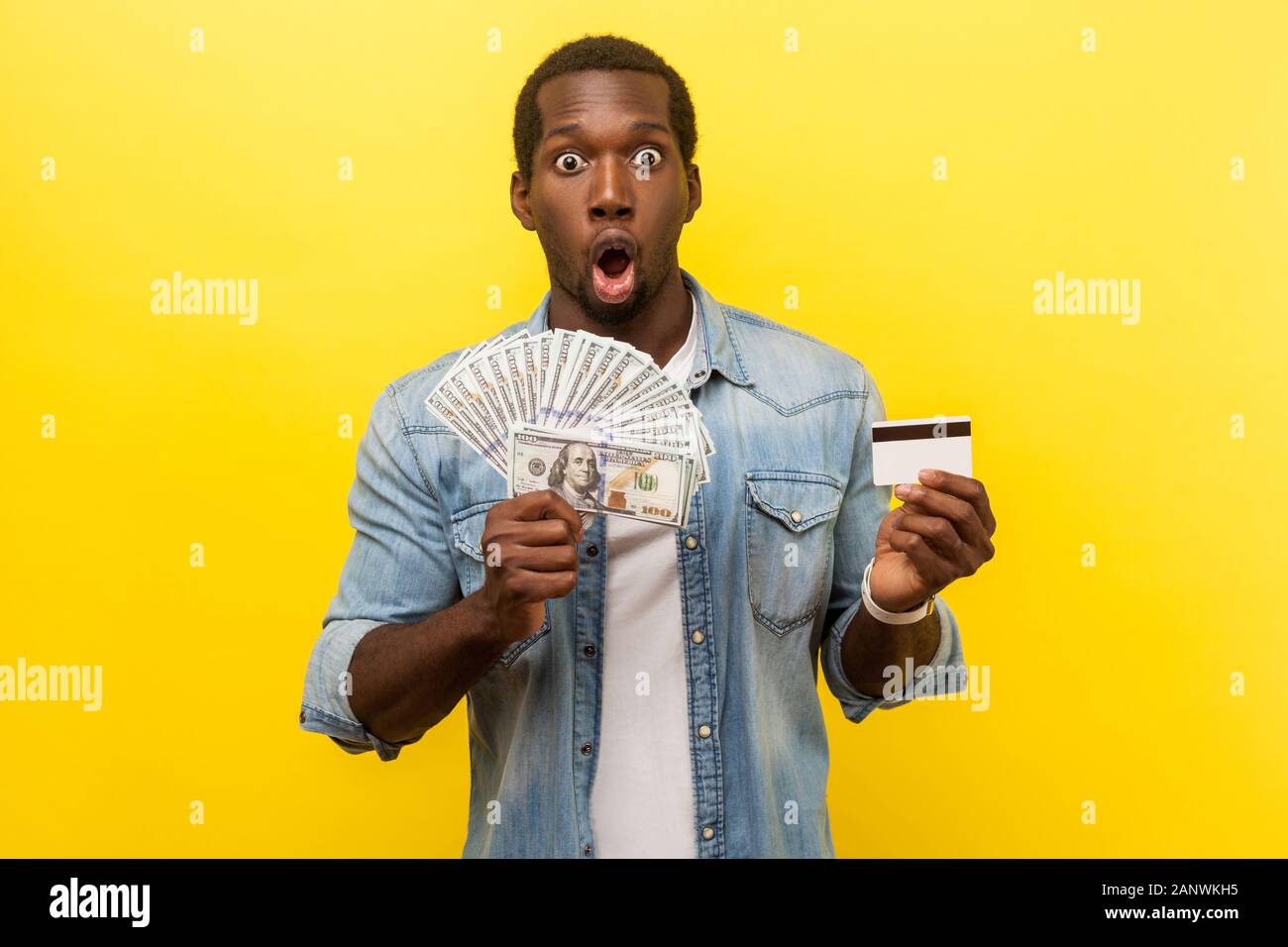 Portrait de la clientèle banque étonné, jeune homme en chemise en jean holding dollar bills et de carte de crédit avec l'expression étonnée, choquée par remise. indoo Banque D'Images