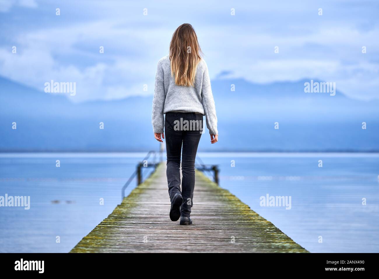 Jeune fille blonde avec de longs cheveux, debout et de retour à l'appareil photo sur une jetée en bois devant un lac calme regardant à l'horizon à Baviera. Voyage Banque D'Images