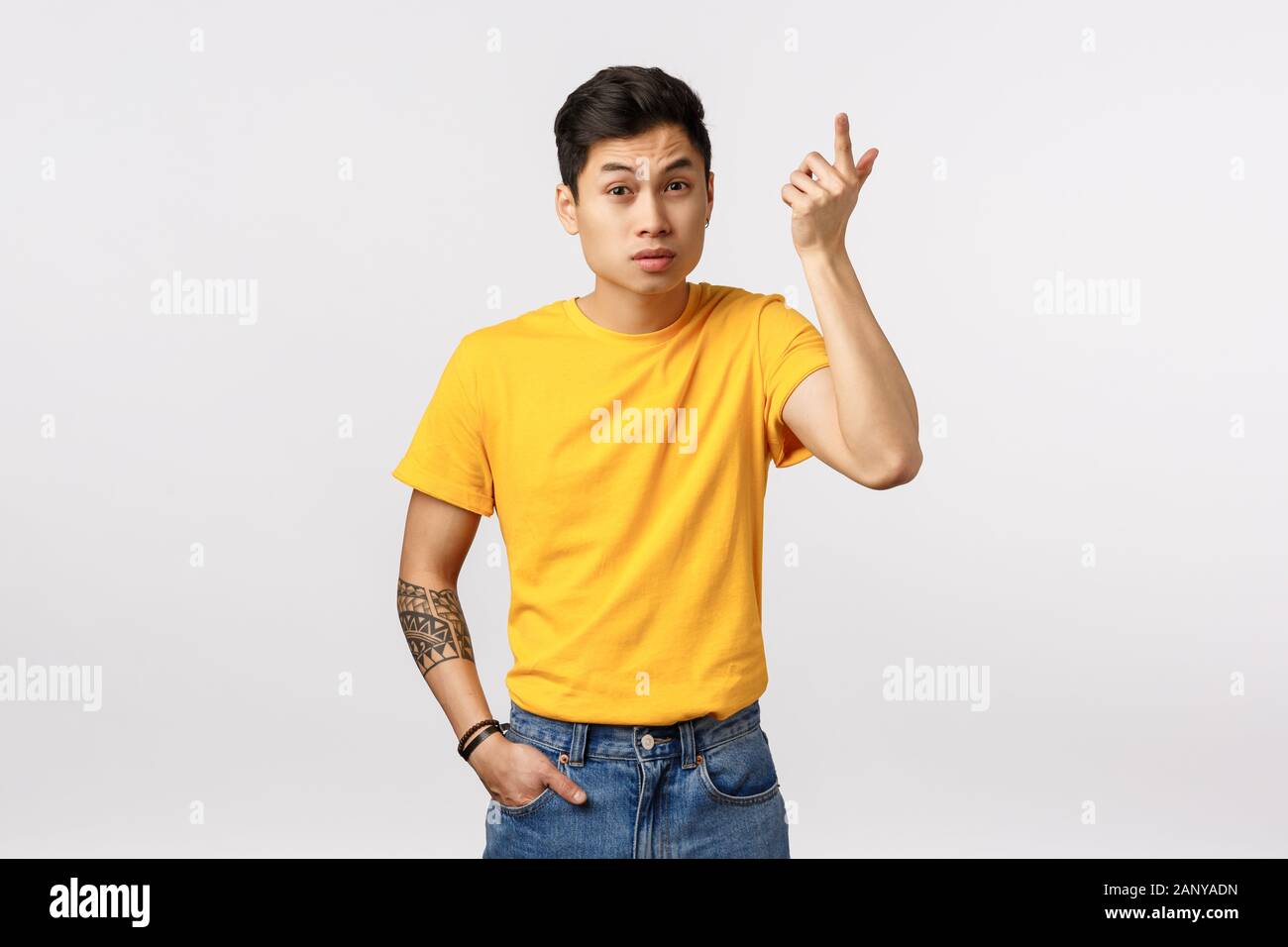 Wtf passe. Jeune homme asiatique ennuyé en t-shirt jaune, de tatouages, de l'index dans la consternation et l'irritation, soulevant les sourcils comme audience Banque D'Images