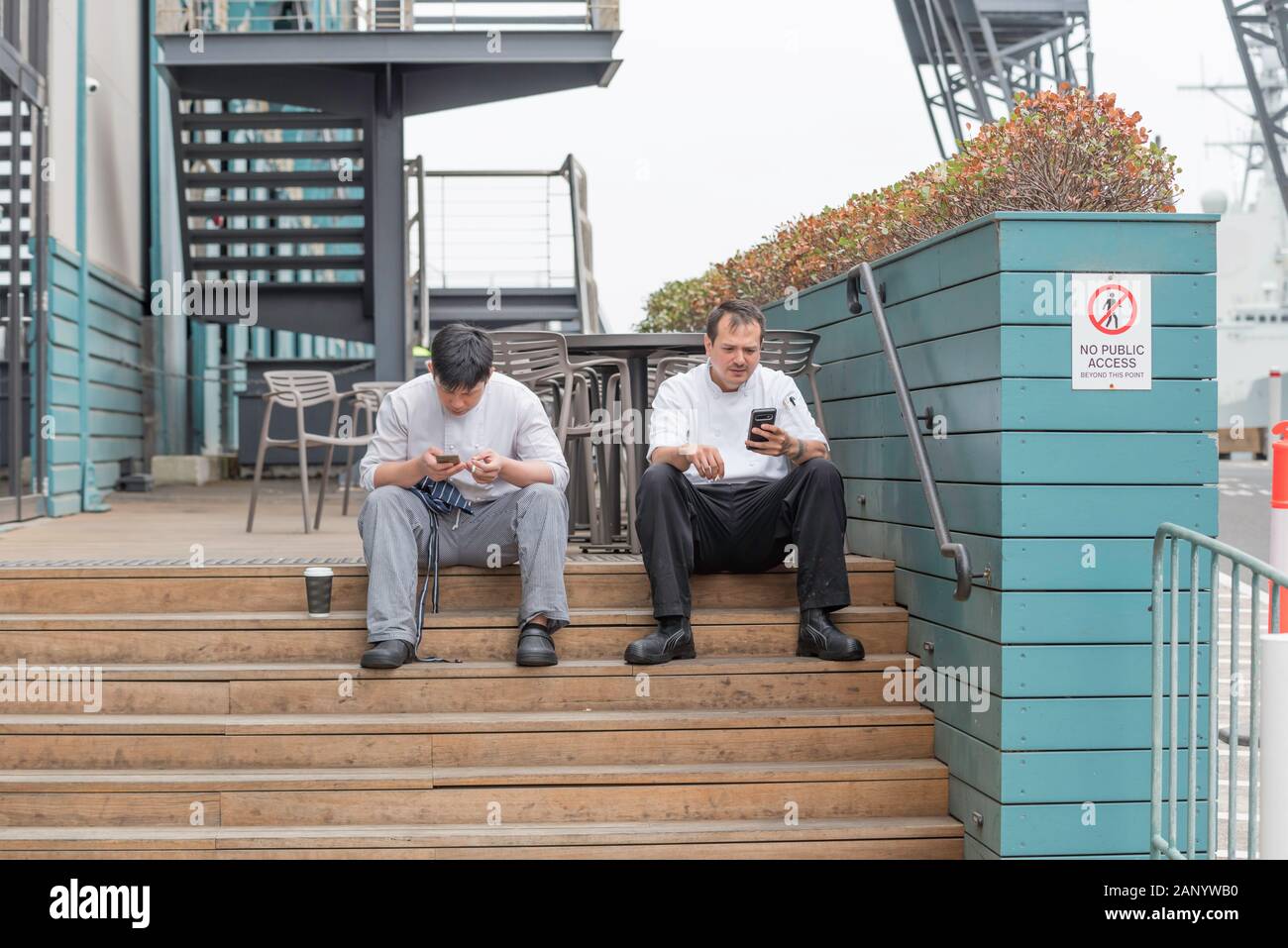 Deux chefs s'assoient à l'extérieur d'un restaurant dans la banlieue de Sydney de Woolloomooloo et fument tout en regardant leurs téléphones mobiles Banque D'Images