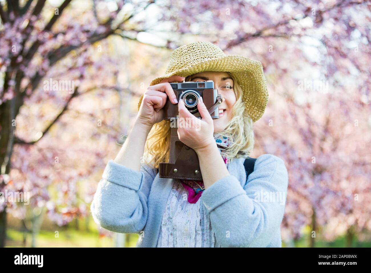 Portrait d'une belle femme dans un chapeau de paille voyageant dans un beau parc avec des cerisiers en fleurs, prenant des photos sur un appareil photo rétro. Tourisme avec sac à dos Banque D'Images