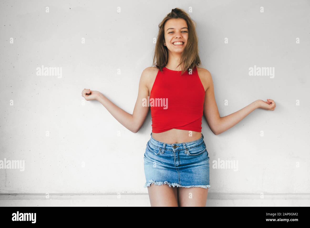 Belle teen girl in red top court et mini jupe en jean riant contre un mur en béton à bras ouverts Banque D'Images