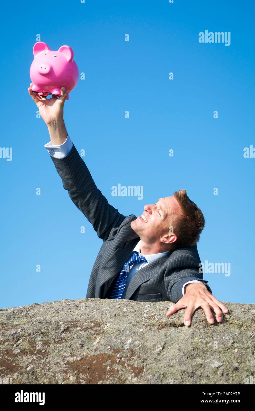 Smiling businessman holding sur un fidèle pink piggy bank rock comme ce qu'il atteint le sommet d'une falaise rocheuse Banque D'Images