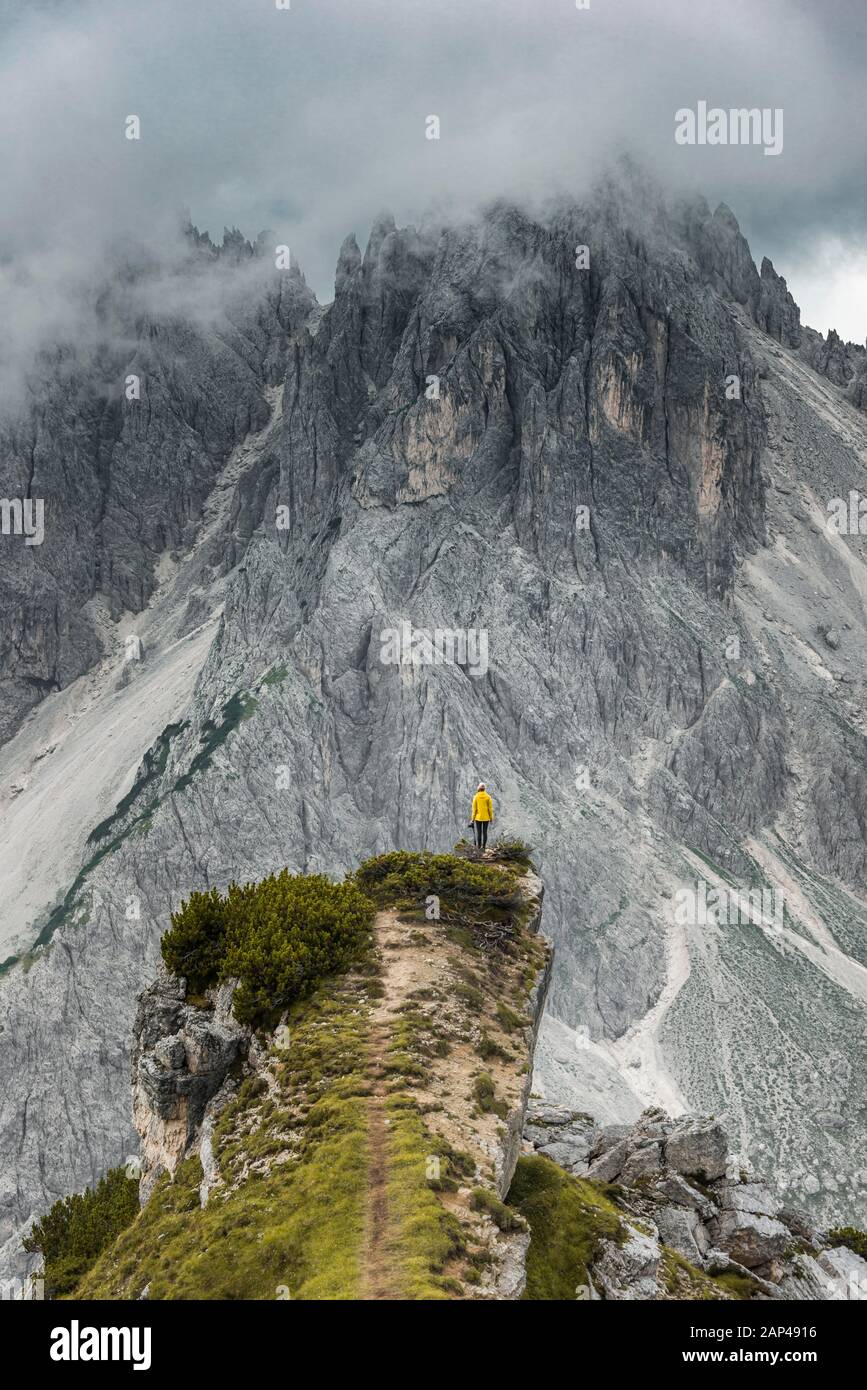 Femme en veste jaune debout sur une crête, derrière ses pics de montagne et ses pics rocheux vifs, ciel nuageux, Cimon le groupe Croda Liscia et Cadini Banque D'Images